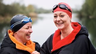 Two women laughing and smiling wearing swim caps, dry robes and goggles next to lake after learning benefits of swimming
