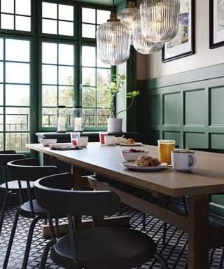 Emerald green dining room with painted wall panels and window frames, large rectangular table, black chairs, artwork, glass pendant lights, black and white floo