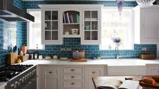 A blue tiled kitchen with bright windows, a marble countertop and white cabinets, a white sink, a wooden island and a black hob with black extractor fans.