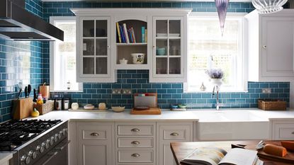 A blue tiled kitchen with bright windows, a marble countertop and white cabinets, a white sink, a wooden island and a black hob with black extractor fans.