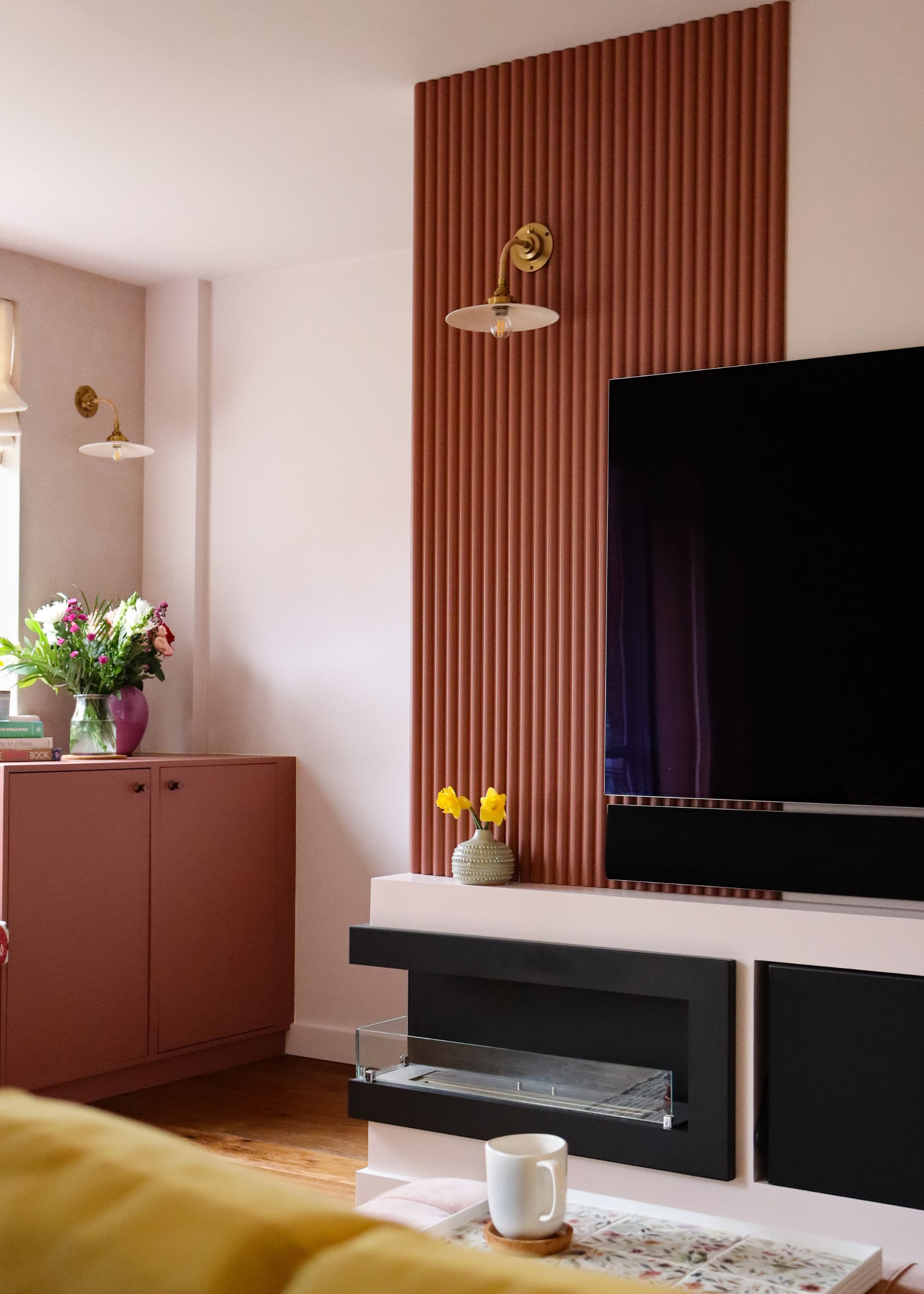 A living room with a tv and panelled walls. On the panelled walls in a lamp. Below the TV is a shelf with extra storage. On the left side of the room is a two door cabinet and on top there are two flowers inside a vase.