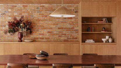 A bright open dining room with a brick wall, a wood cabinet, a woven pendant, and a wood dining set with a bowl of aubergines and a vase of flowers