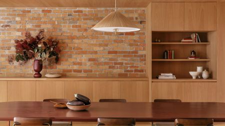 A bright open dining room with a brick wall, a wood cabinet, a woven pendant, and a wood dining set with a bowl of aubergines and a vase of flowers