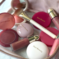 A close up shot of a selection of the various Rare Beauty blushers and a brush within a cosmetic bag