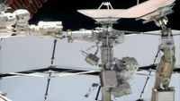 two astronauts dressed in white spacesuits work near a couple of antenna dishes outside of a space station.