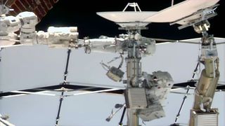 two astronauts dressed in white spacesuits work near a couple of antenna dishes outside of a space station.