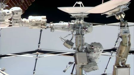 two astronauts dressed in white spacesuits work near a couple of antenna dishes outside of a space station.