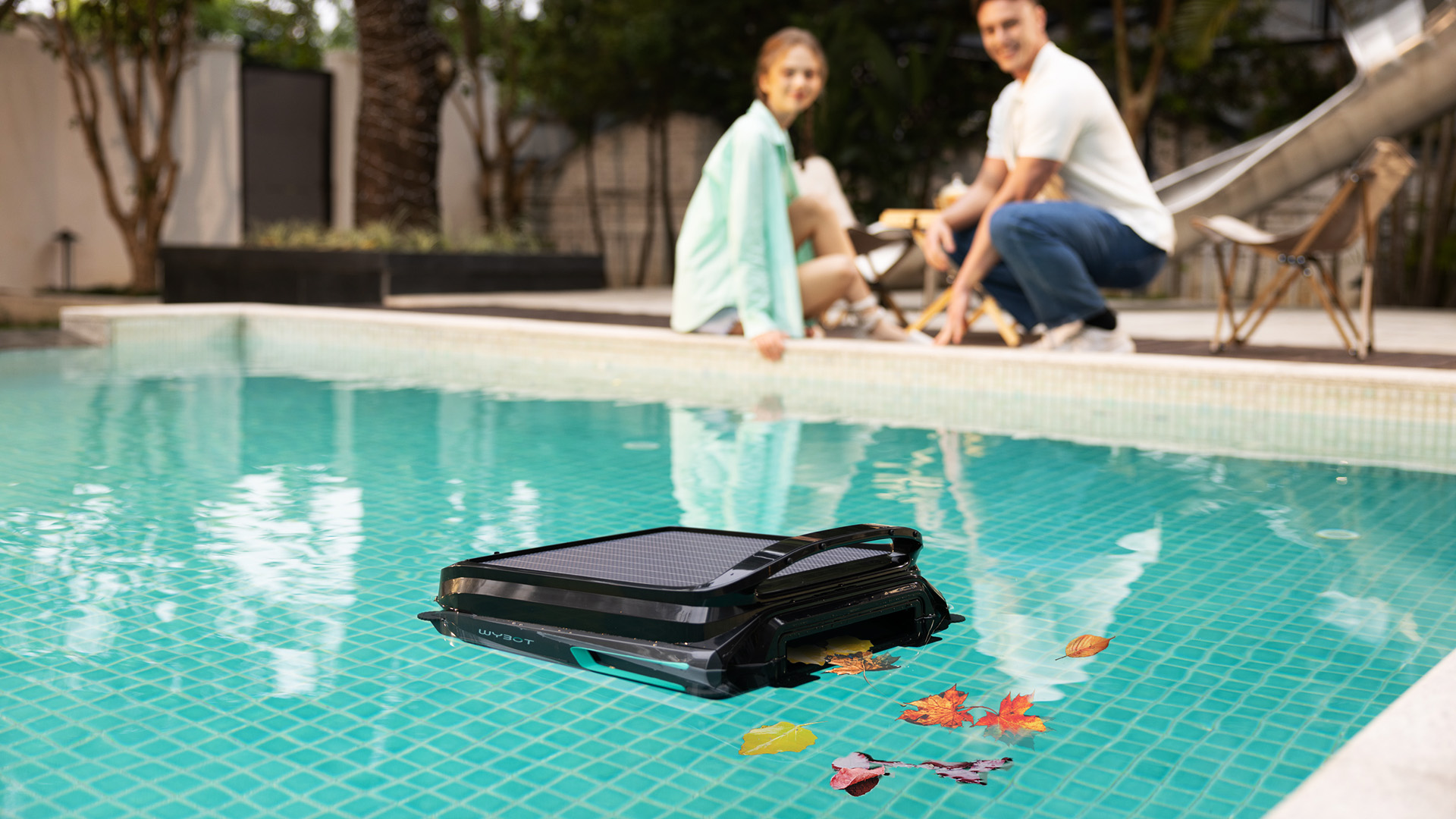 A WYBOT F1 pool skimmer clears the water surface of debris in a large mosaic tiled pool while a family sits in the background.