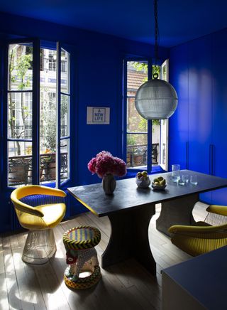 Image of a bright, indigo-blue dining room with two large french windows. The dining room is a slate stone material with yellow basket dining chairs. There is a globe pendant light hanging above the dining table and a vase of pink flowers resting on the table.