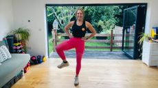 Fitness journalist Maddy Biddulph stands performing a hip opening exercise in her living room. Her hands are on her hips and her right knee is bent and held out to the side. She is smiling and behind her we see open glass doors and a green leafy garden.