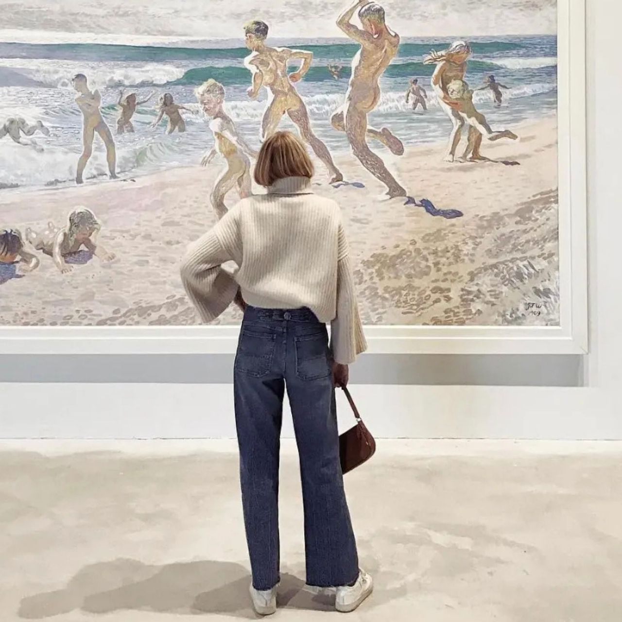 A woman looks at a painting of kids playing at the beach while wearing a white sweater and jeans