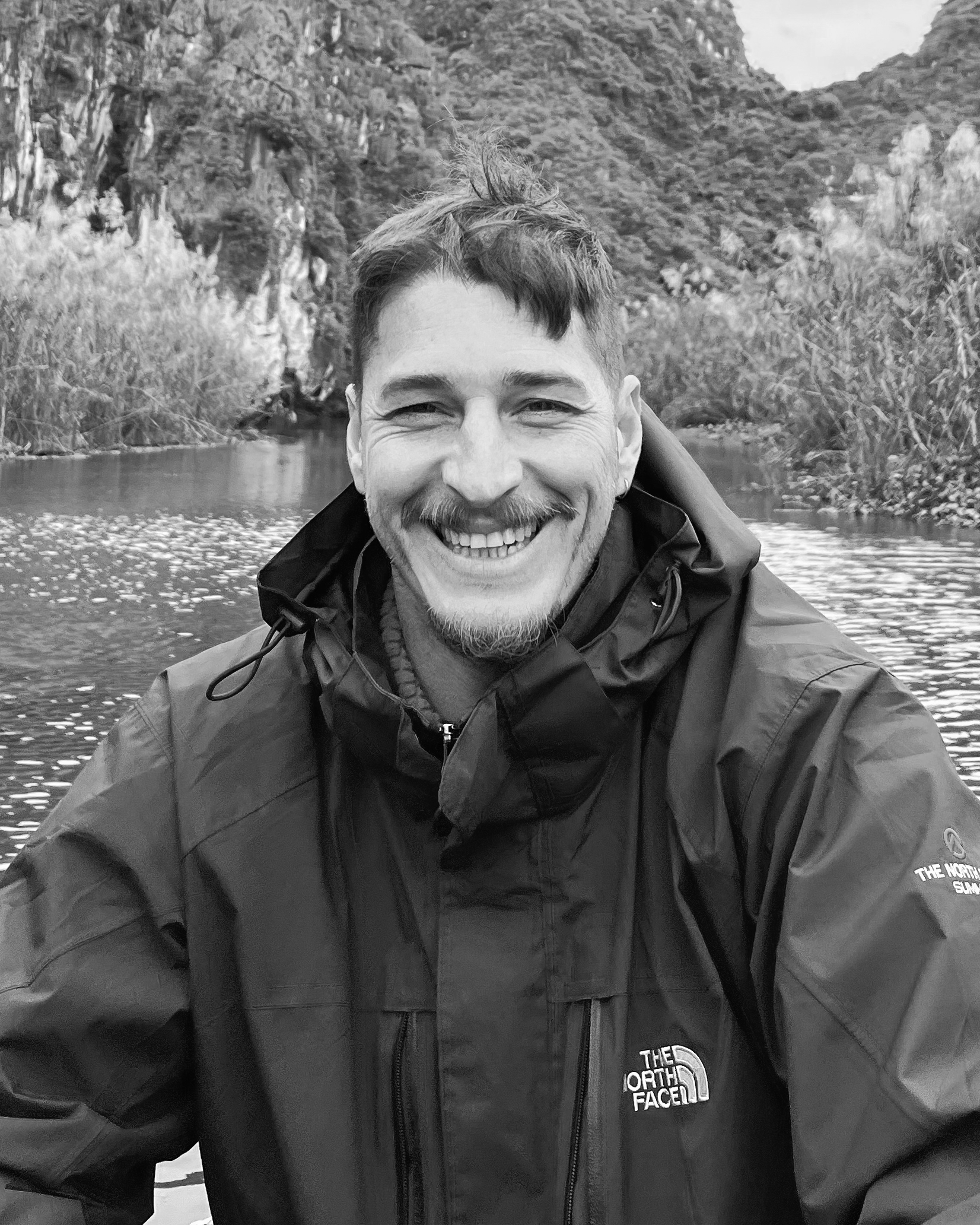 A person wearing a black North Face jacket sits by a calm river surrounded by lush greenery and rocky cliffs in a monochromatic setting