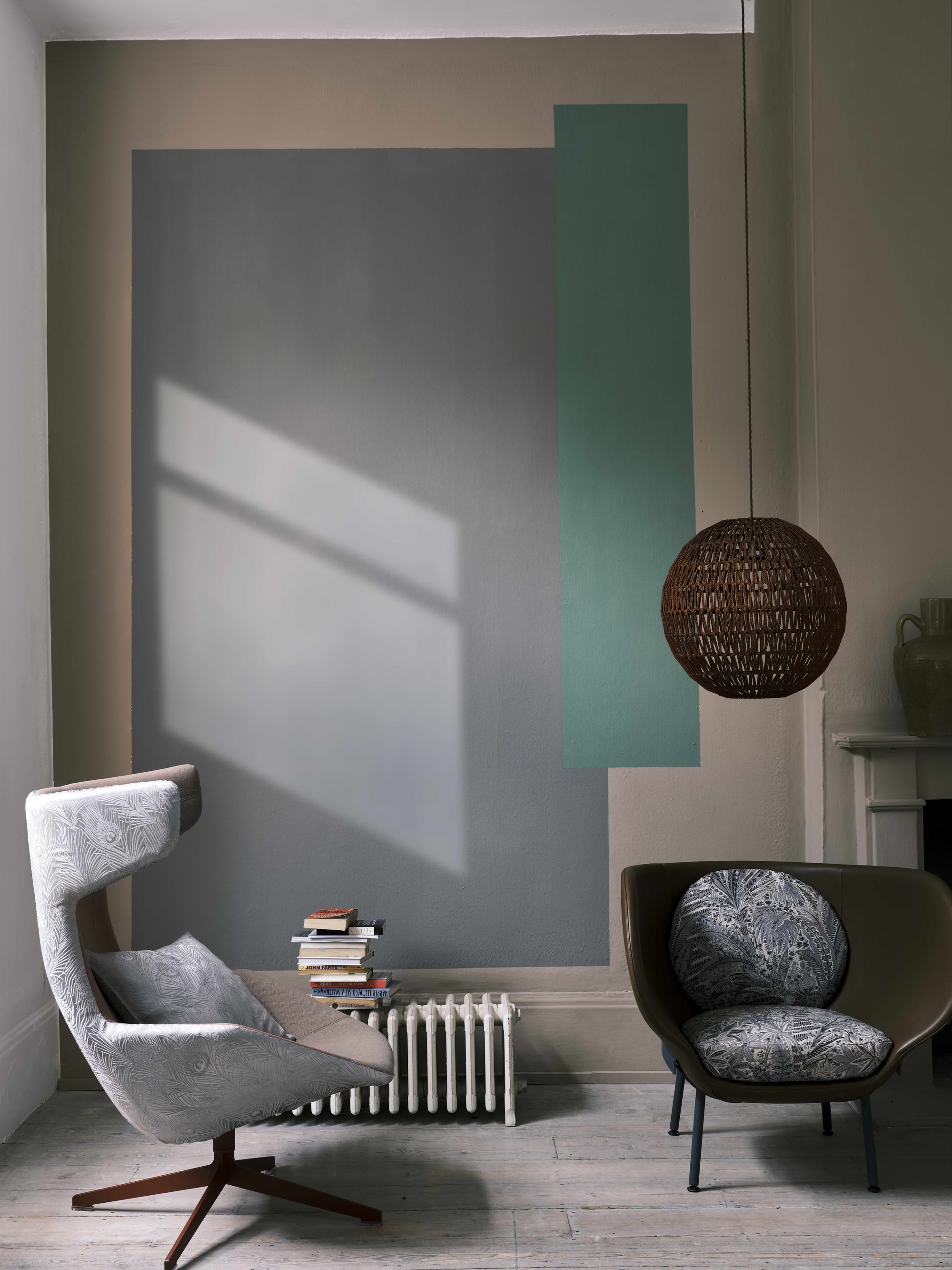 living room corner with gray armchairs, short white radiator with a stack of books on top, and a beige wall painted with a gray and green graphic, and a rattan pendant light hanging above
