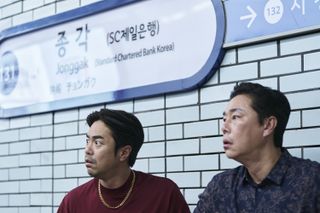 Two men sit in front of a subway walk covered in white tiles, under a sign that reads "Jonggak Standard Chartered Bank Korea," in 'Squid Game' season 2.