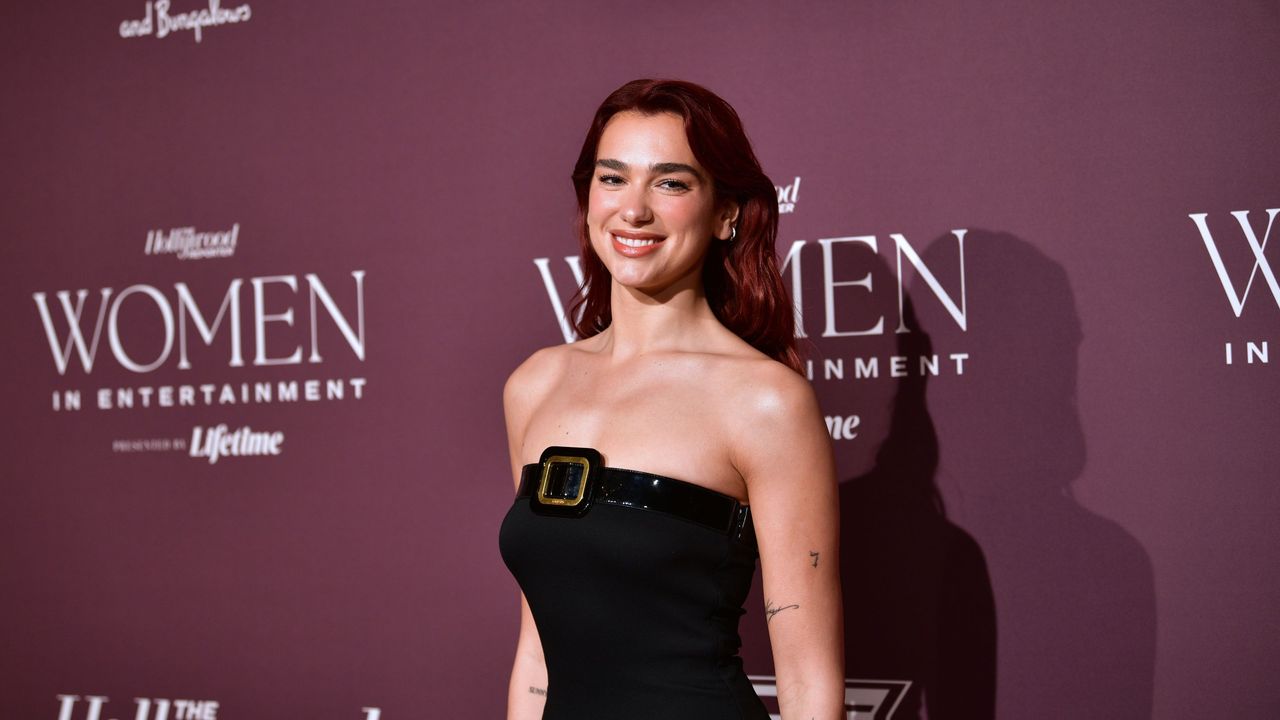 Dua Lipa&#039;s quiet luxury kitchen is so chic. Here is the singer wearing a black dress, standing against a purple background with white lettering