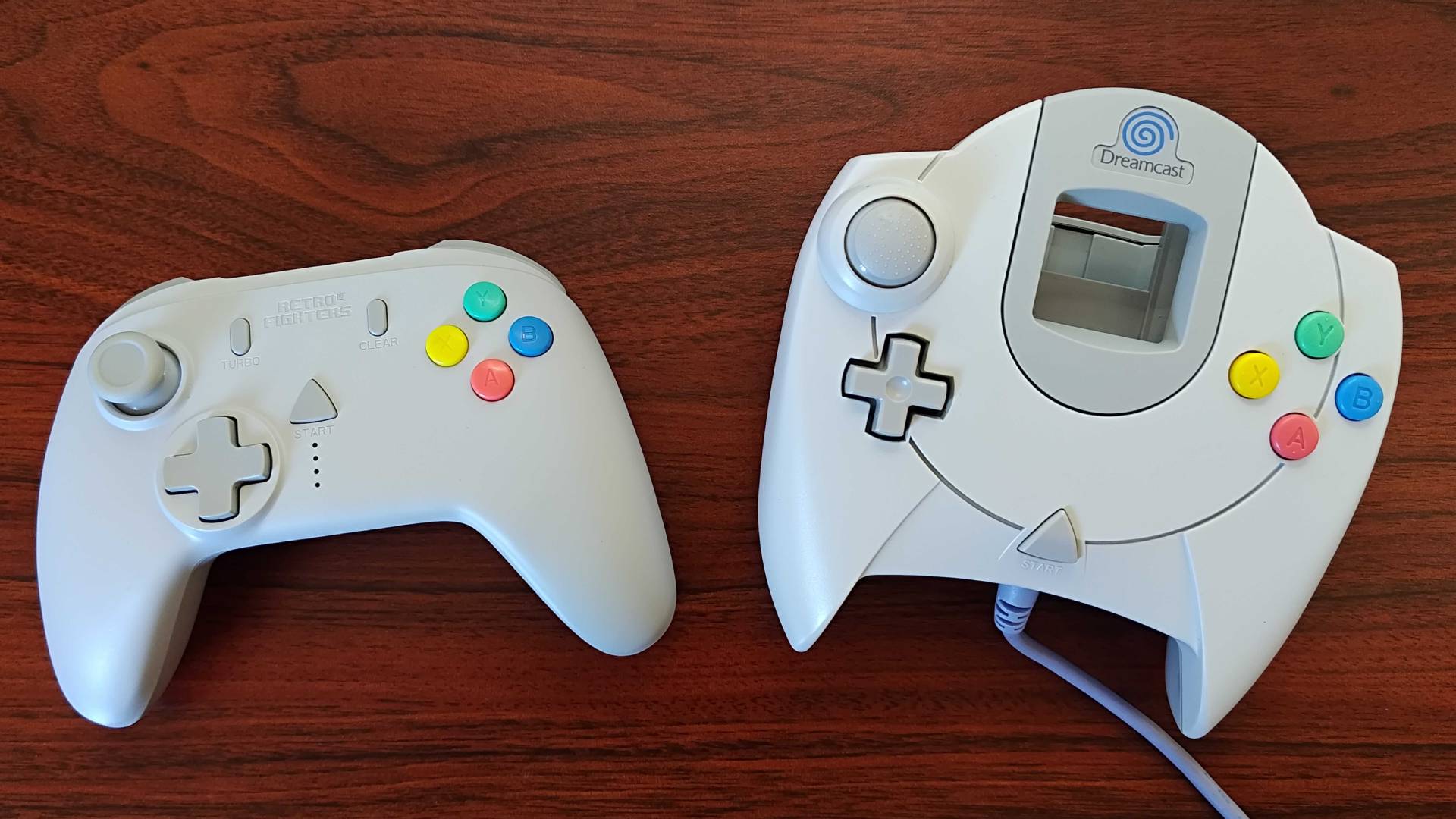 Retro Fighters StrikerDC on left and original Sega Dreamcast controller on right on woodgrain table.