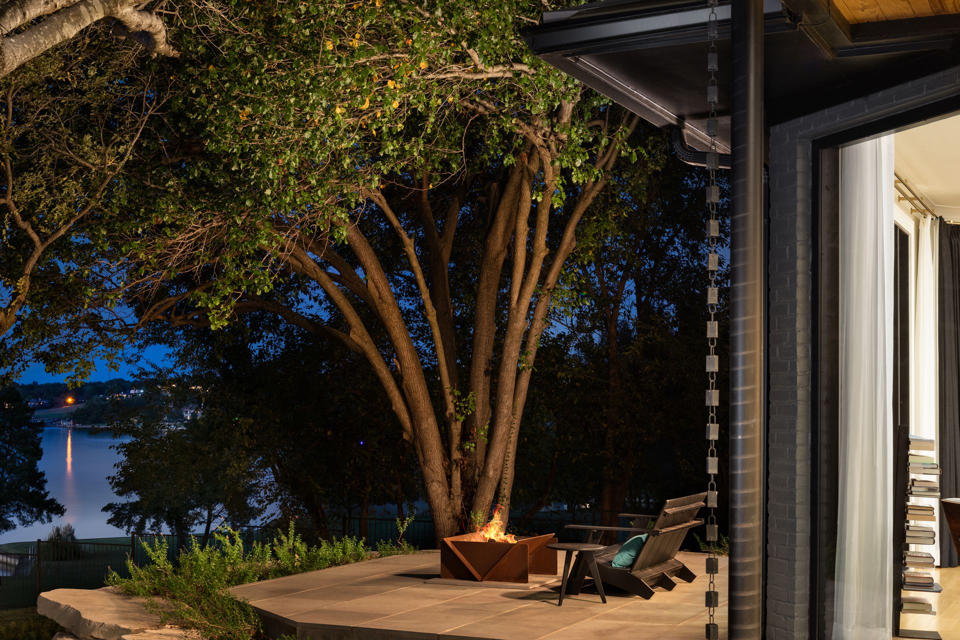 An outdoor space with a lit firepit, black armchair and side table in front of a large tree looking out onto a lake at nighttime