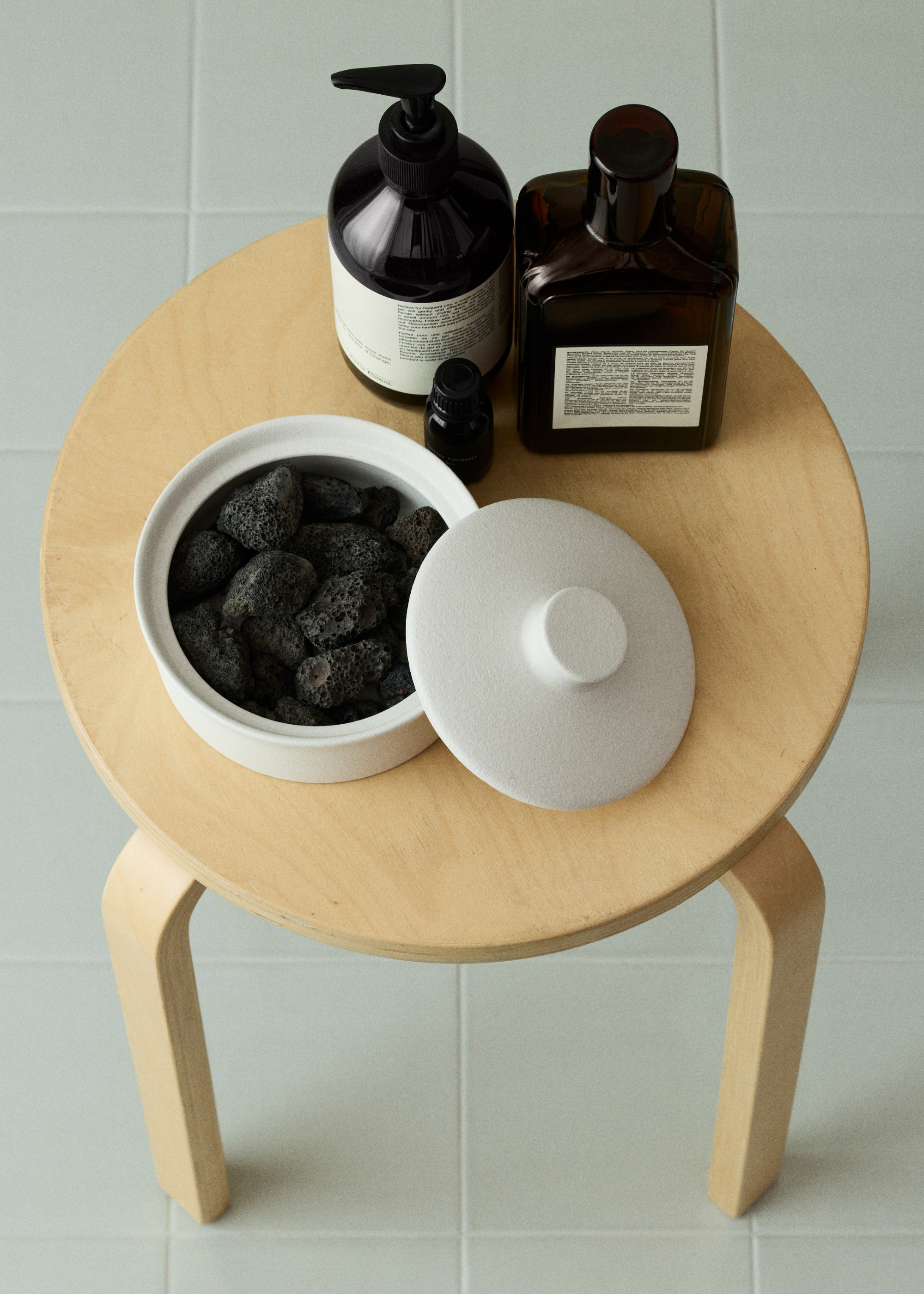 A wooden stool with a lava rock diffuser in a white vessel and a bottle of essential oils beside a hand soap and a lotion on a light tiled floor