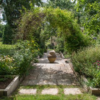 a long paved garden path with raised beds filled with plants and flowers and an arch made from climbing plants