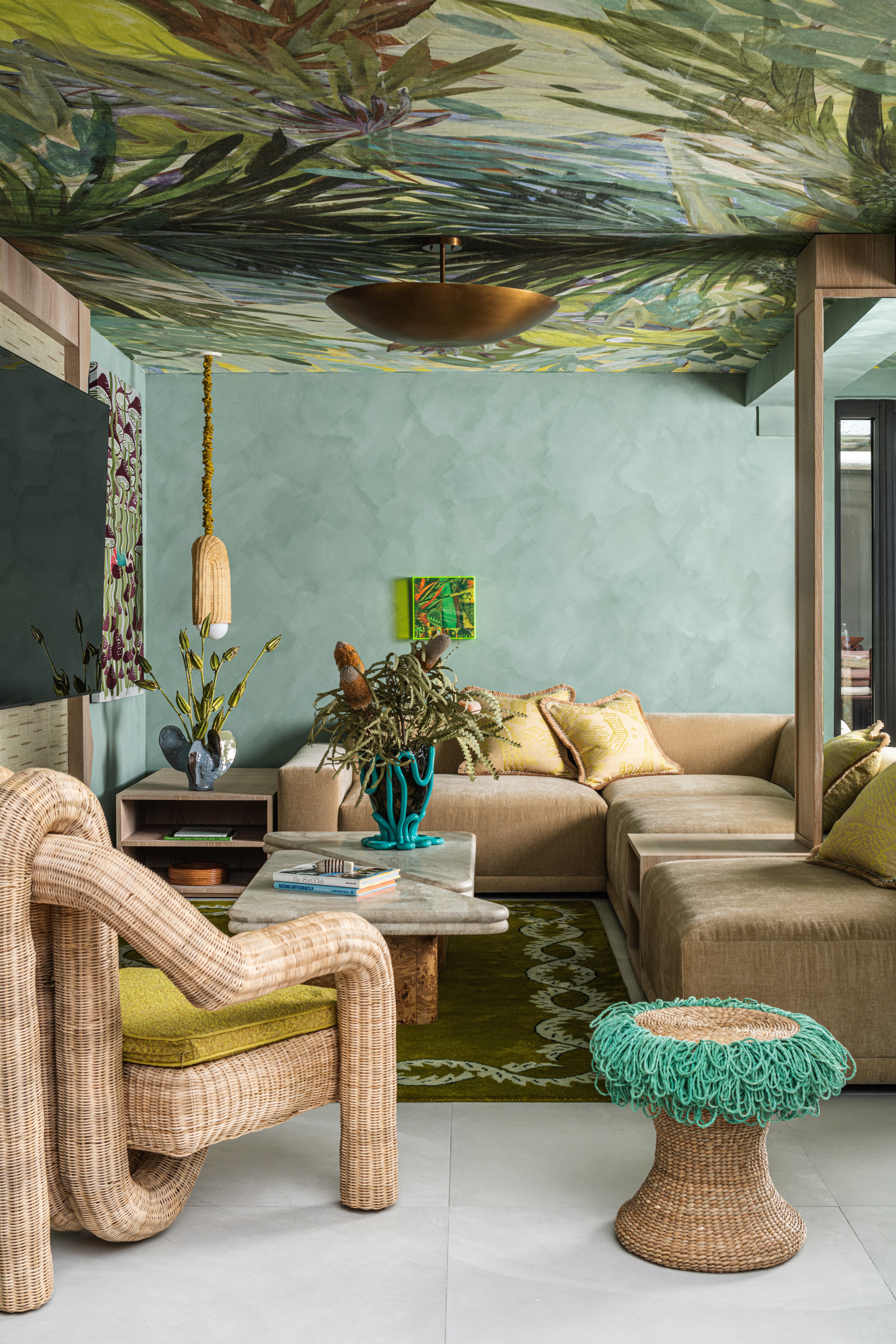Interior of a tropical home with teal walls, zellige tiles surfaces, wavy, sand-coated details, rattan furniture mixed with bright aquamarine and ochre touches, and a botanical-themed mural ceiling.