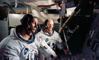 two men in white spacesuits look at dials and knobs on a control panel