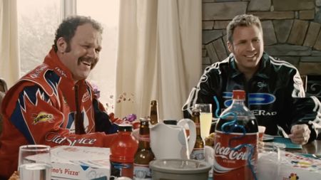 John C. Reilly and Will Ferrell are shown in Talladega Nights, wearing racing gear and enjoying a table full of food. 