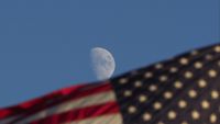 A photo showing the moon in a blue sky. On the lower half of the screen, there's an American flag.