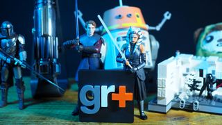 Action figures, lightsaber hilts, droid animatronics, plushies, and Lego sets from Star Wars on a wooden table in front of a GR+ logo