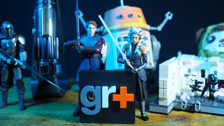Action figures, lightsaber hilts, droid animatronics, plushies, and Lego sets from Star Wars on a wooden table in front of a GR+ logo