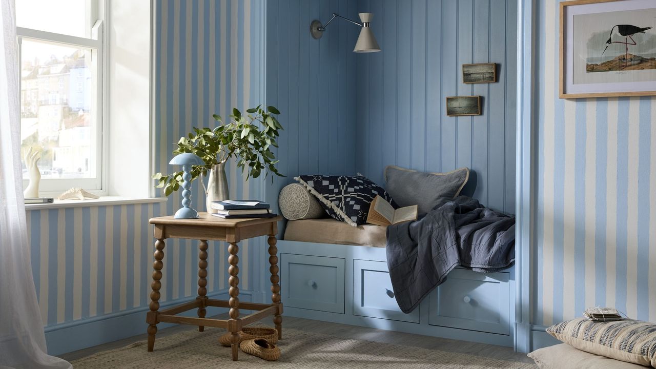 Image of a daybed book painted light blue in a room that has blue and white stripped walls. There is a wooden bobbin side table next to the daybed with a light blue lamp and vase on it.
