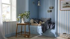 Image of a daybed book painted light blue in a room that has blue and white stripped walls. There is a wooden bobbin side table next to the daybed with a light blue lamp and vase on it.