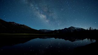 Star-studded night sky above trees and rolling hills, the starlight is reflected in a seasonal pond below. 