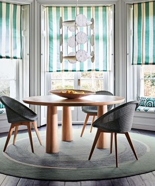 Round dining table with conical legs and black tub dining chairs on circular green rug, green and white striped Roman blinds.