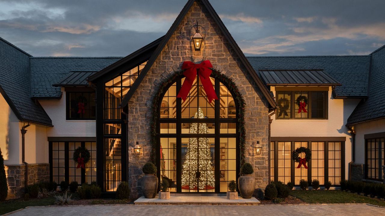 large stone home with a pitched roof and large arched glass window showing a tall lit Christmas tree with a large row bow hung from the pitched roof and wreaths on the windows