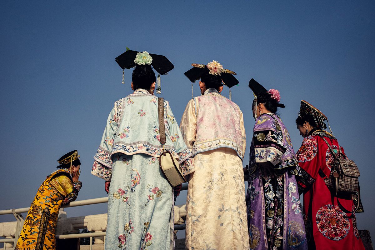 Five women in traditional colorful attire stand outdoors, gazing at something in the distance against a clear blue sky