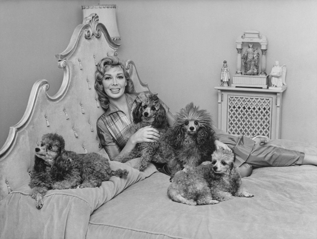 A glamorous woman reclines on an ornate bed beside four poodles, each curled comfortably around her, in a 1950s interior setting.