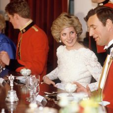 Princess Diana wearing a white lace gown sitting at a banquet table between two soldiers in red uniforms