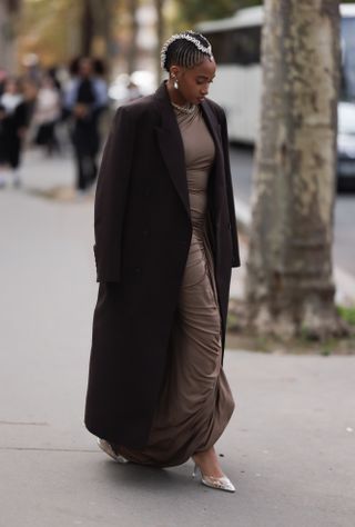 A Fashion Week guest is seen wearing braided hair with shells in it, silver drop earrings, tiny gold hoop earrings, and a gold multi-row ear cuff, a chunky chain necklace, an oversized ankle-length dark brown coat with notch lapel, underneath a floor-length brown fitted dress with high round neckline and gathered hip detail, and pointed transparent heeled mules with rhinestone embellishments before the Atlein Womenswear Spring/Summer 2025 Show as part of Paris Fashion Week on September 29, 2024 in Paris, France