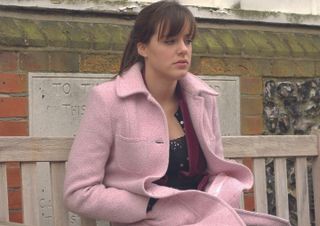 Zoe Slater sitting on a bench wearing a pink coat