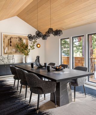 Large dining room with wooden ceiling paneling, large rectangular marble dining table with six upholstered chairs, black and cream carpet, black metallic cluster pendant, black low sideboard with framed artwork and vase of flowers
