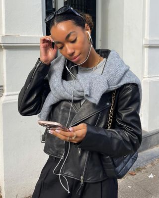 Dutch fashion influencer Amaka Hamelijnck with wired Apple earbuds, a gray sweater over her shoulder, and a black leather jacket