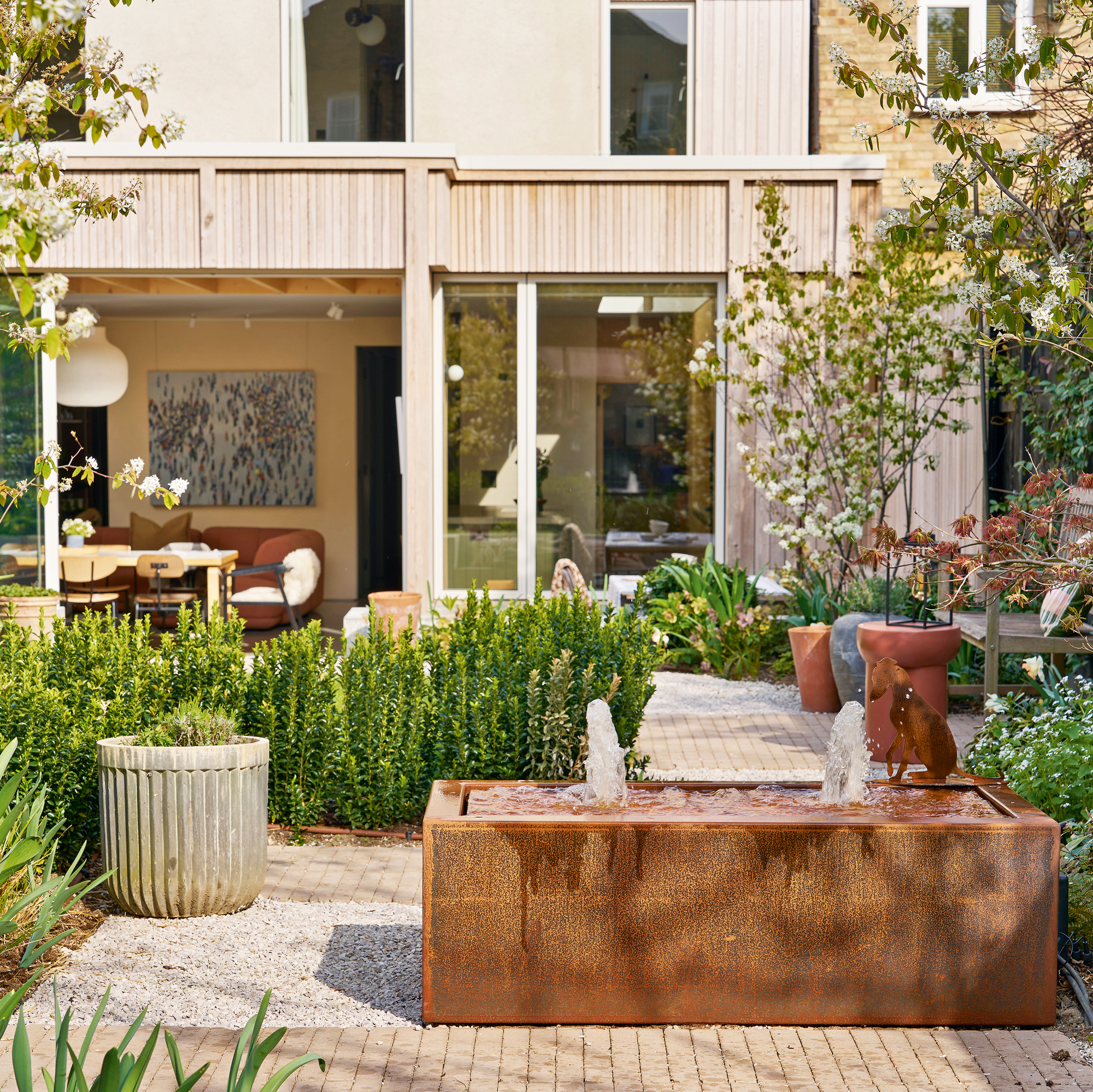 the rear of a property with a large garden with a corten steel water feature, brick paving and plenty of trees and planting