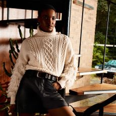 Model wearing a white jumper with tassels on the arms and black shorts sold at Oasis leaning on a spiral staircase 