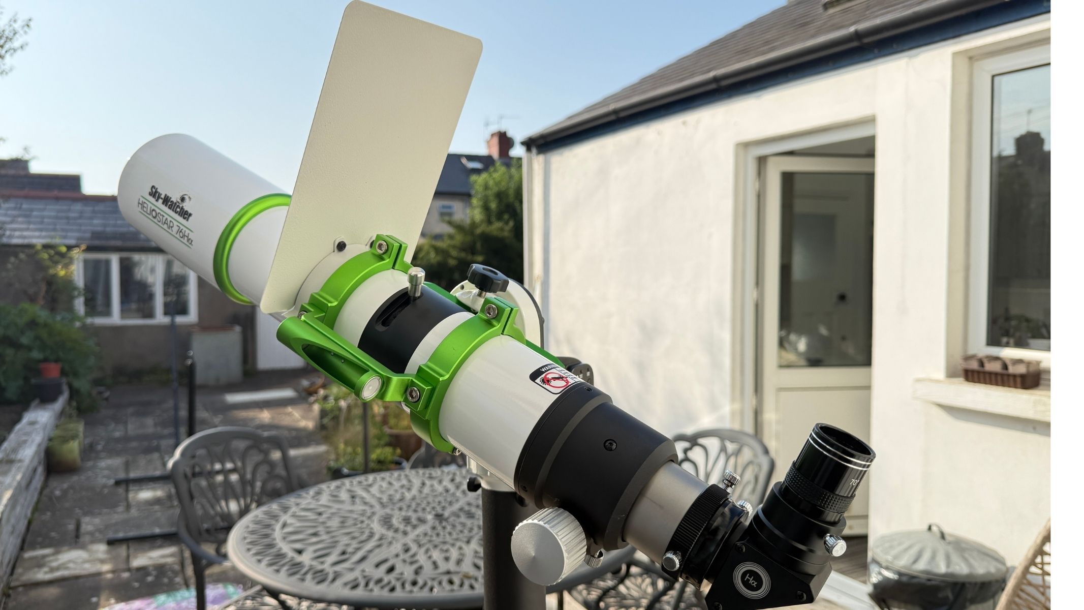 The Sky-Watcher Heliostar 76mm H-alpha solar telescope with clip-on sunshade attached with a house and table in the background.