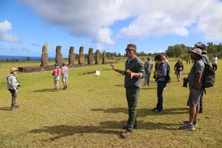 several people stand around looking at seven large statues, a man in the center of the image is explaining something with his hands outstretched.