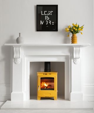 yellow log burning stove in white fireplace in room with white walls, on fireplace are yellow vase and yellow flowers, grey vase and black and white typography print saying less is more