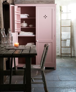 laundry room with stone floor, brick walls, table and chair, pink linen cupboard with folded items, ladder storage near window