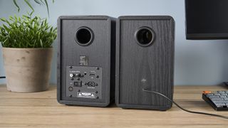 a pair of black wood speakers with a yellow kevlar cone photographed on a wood desk with a computer and blue wall and a plant in shot