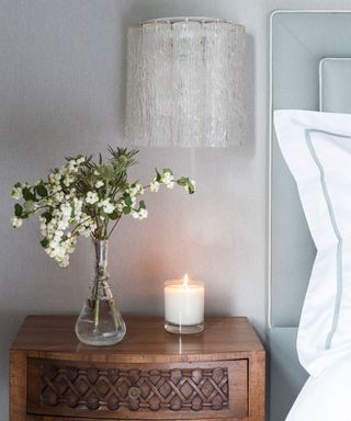 Luxury bedroom ideas illustrated by a candle and vase of flowers on a dark wood bedside table.