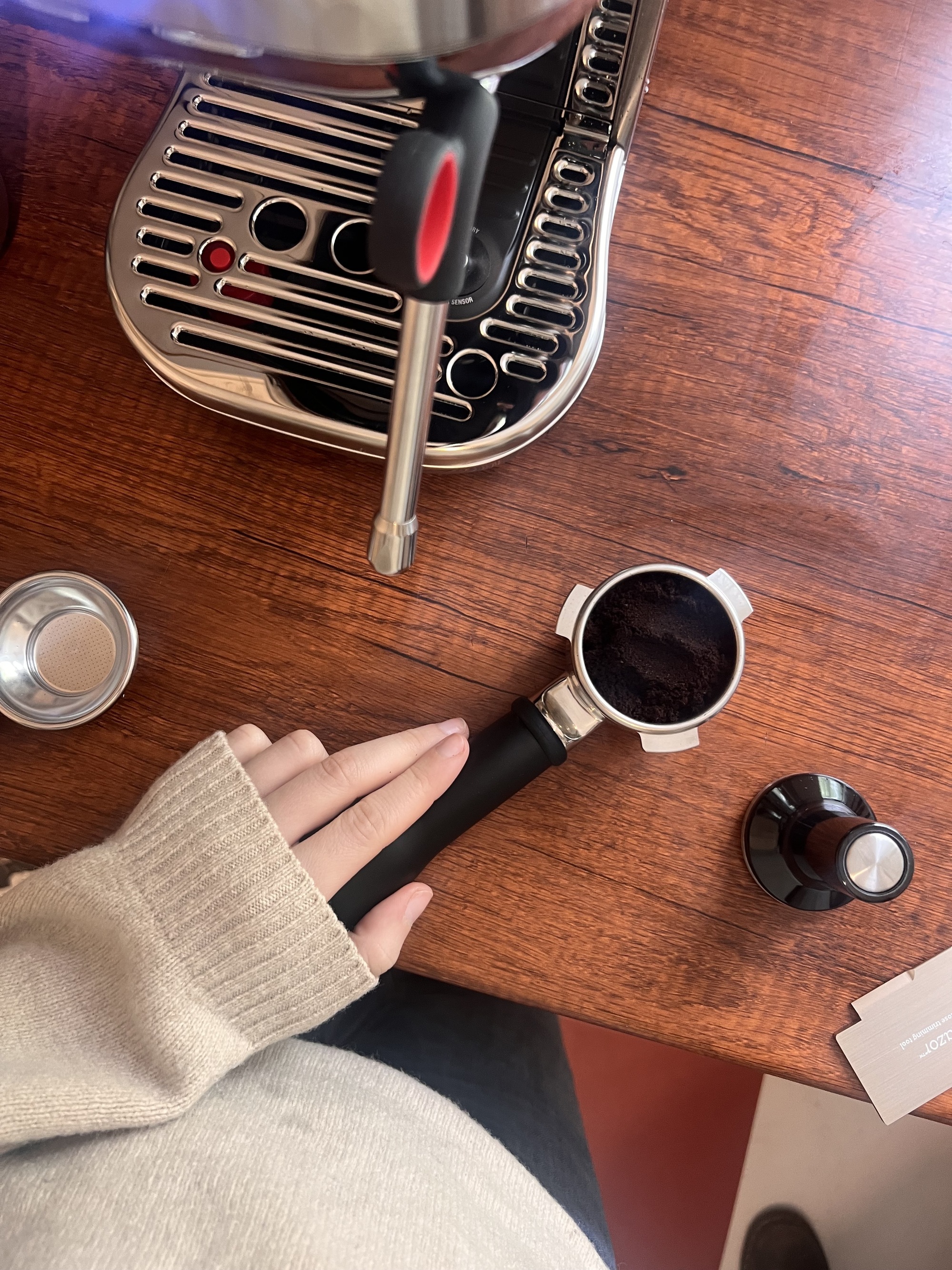 Image of a hand in a beige sweater holding a portafilter filled with coffee grounds on a wooden countertop with a Sage Bambino Plus machine in the corner